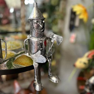 May include: A silver-colored metal figurine of the Tin Man from The Wizard of Oz. The figurine is seated with an axe in one hand and a heart-shaped detail on its chest. The figure has a conical hat and a smiling face.