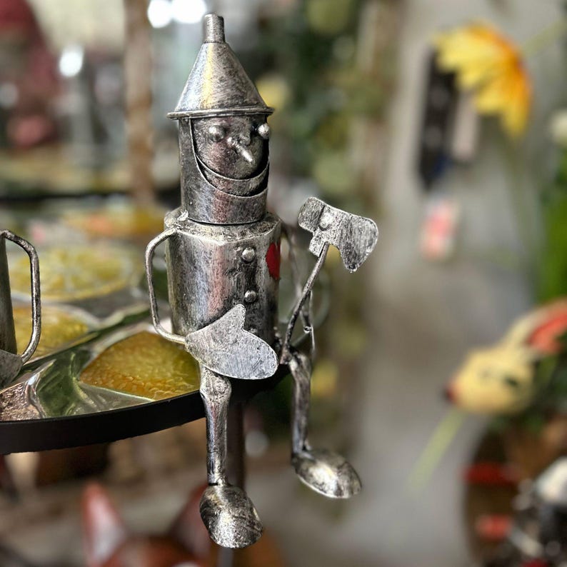 May include: A silver-colored metal figurine of the Tin Man from The Wizard of Oz. The figurine is seated with an axe in one hand and a heart-shaped detail on its chest. The figure has a conical hat and a smiling face.