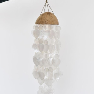 May include: A decorative wind chime with a natural coconut shell top and strands of heart-shaped, translucent shell pieces. The chime is suspended by a woven cord, creating an airy aesthetic. The color palette is neutral, with a focus on natural materials and textures.