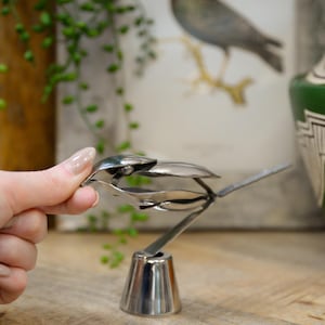 May include: A handcrafted, silver-colored metal bird sculpture. The bird is made from spoons and forks, with a detailed beak, wings, and tail. The sculpture is mounted on a conical base, and is displayed on a wooden surface.