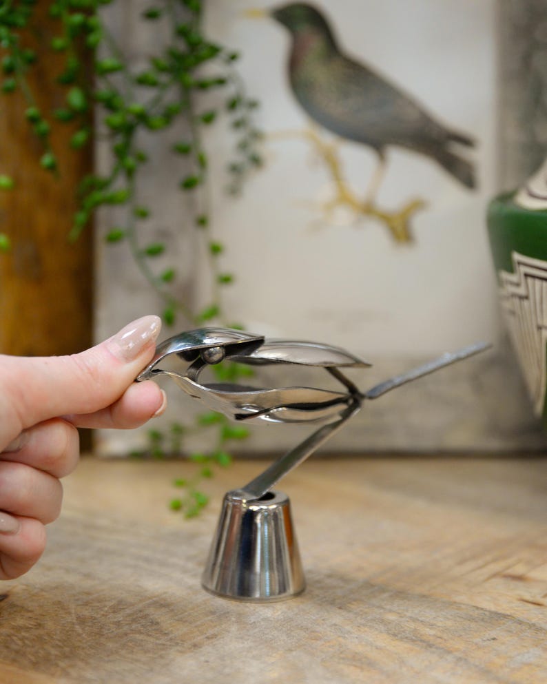 May include: A handcrafted, silver-colored metal bird sculpture. The bird is made from spoons and forks, with a detailed beak, wings, and tail. The sculpture is mounted on a conical base, and is displayed on a wooden surface.