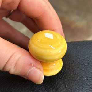 May include: A round, glossy cabinet knob with a smooth, bright yellow finish and subtle, swirling tonal variations. The knob features a classic mushroom-shaped profile with a rounded top and a tapered base, designed for furniture or cabinetry hardware.