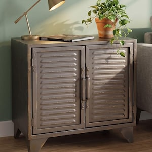 May include: A dark grey metal cabinet with two louvered doors and vertical handles. The cabinet features a distressed finish with visible wear along the edges and stands on four angled legs. The top surface is flat and rectangular.
