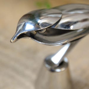 May include: A polished silver bird sculpture with a sleek, modern design. The bird's head is detailed with a pointed beak and a smooth, reflective surface. The sculpture is mounted on a conical base, creating a striking decorative piece.