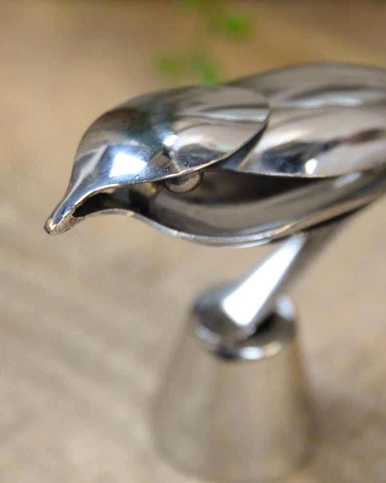 May include: A polished silver bird sculpture with a sleek, modern design. The bird's head is detailed with a pointed beak and a smooth, reflective surface. The sculpture is mounted on a conical base, creating a striking decorative piece.