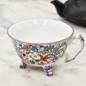 May include: White teacup with a floral design in pink, orange, blue, and purple. The teacup has a purple rim and is supported by three small feet. A black teapot is in the background.