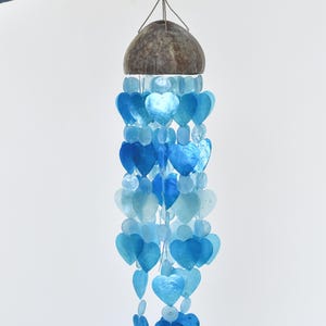May include: A decorative wind chime featuring a coconut shell top and strands of blue heart-shaped shells. The shells are in varying shades of blue. The chime is suspended by a natural fibre cord.
