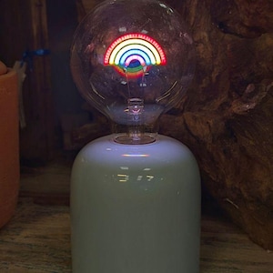 May include: A clear glass globe light bulb featuring an internal glowing rainbow filament, set into a smooth, cylindrical white ceramic base. The rainbow design displays vibrant red, orange, yellow, green, blue, and violet arcs of light.
