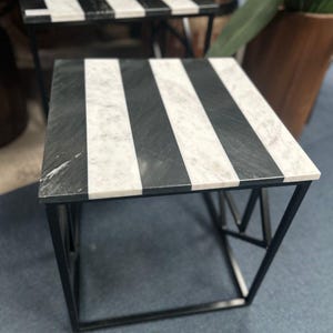 May include: A square side table with a black metal frame and a tabletop featuring alternating black and white stripes. The table's design is modern and geometric, suitable for various interior styles. The table is approximately 46 cm square.