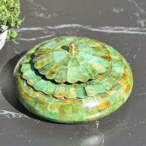 May include: A round, lidded decorative box with a mosaic-like pattern in shades of green and gold. The lid has a scalloped edge and a small, gold-coloured knob. The box is on a dark surface.