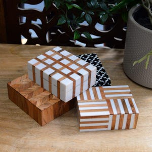 May include: Several decorative wooden boxes with different patterns. One box has a herringbone design, while others display white and brown stripes or a grid. The boxes are displayed on a wooden surface.
