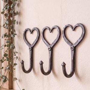 May include: Three dark grey metal wall hooks, each featuring a heart-shaped top and a curved hook at the bottom. The hooks have a textured, hammered finish and are secured with a visible screw at the centre of each heart design.