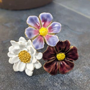May include: A floral brooch with three enamel flowers in white, purple, and burgundy. Each flower has a gold centre and is set against a dark grey background. The brooch is likely made of metal.