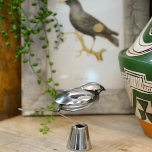 May include: A handcrafted metal bird sculpture made from spoons and other utensils. The bird is perched on a small, silver stand. A framed bird print and a green and white ceramic pot are in the background.