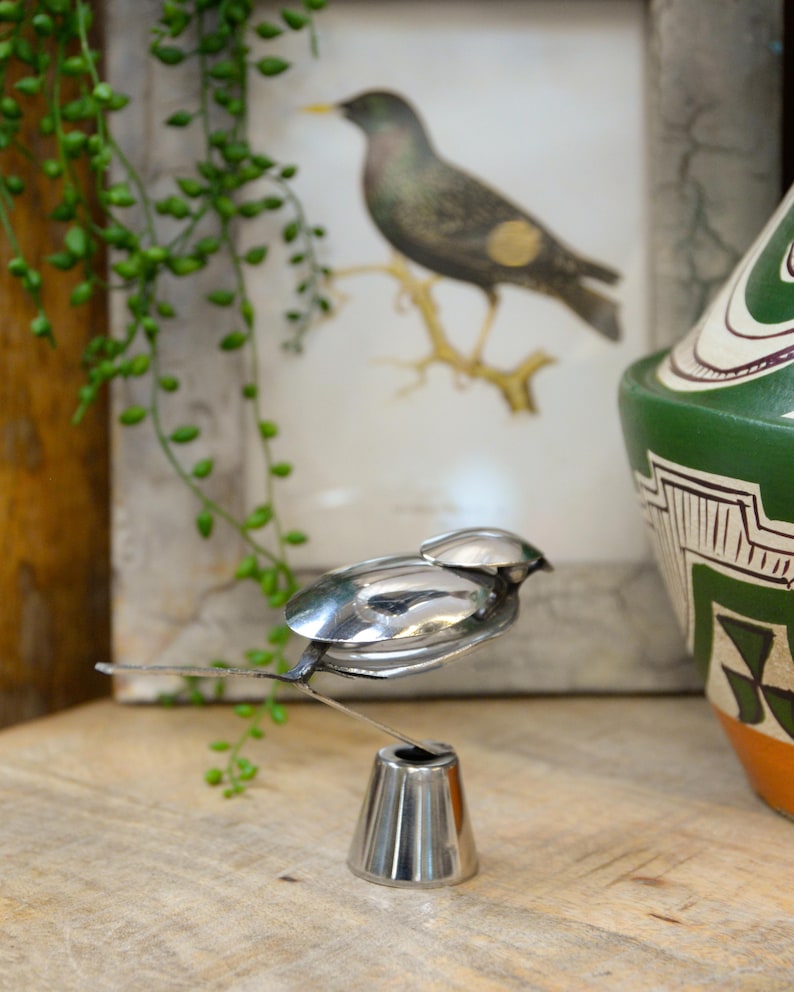 May include: A handcrafted metal bird sculpture made from spoons and other utensils. The bird is perched on a small, silver stand. A framed bird print and a green and white ceramic pot are in the background.