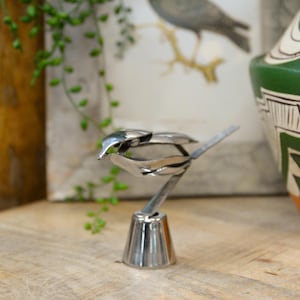 May include: A handcrafted, silver-colored metal bird sculpture. The bird is perched on a decorative base, with its wings and tail artfully crafted. The sculpture is displayed on a wooden surface, with a blurred background featuring a bird illustration and greenery.
