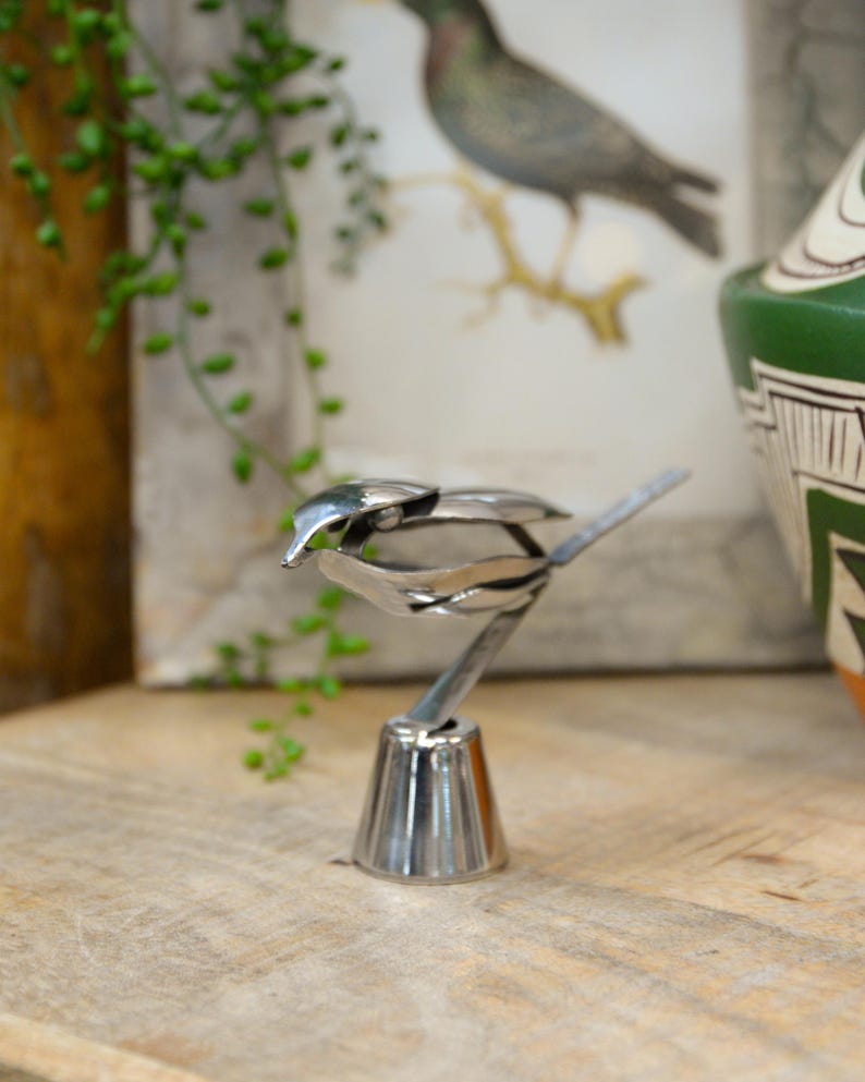 May include: A handcrafted, silver-colored metal bird sculpture. The bird is perched on a decorative base, with its wings and tail artfully crafted. The sculpture is displayed on a wooden surface, with a blurred background featuring a bird illustration and greenery.
