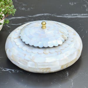 May include: A round, white decorative box with a lid. The box is covered in iridescent, mother-of-pearl tiles. The lid has a scalloped edge and a small, gold-coloured knob. The box is on a dark surface.