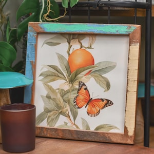 May include: A framed botanical print of an orange and a butterfly. The artwork is set in a rustic wooden frame with blue and green accents. A brown candle holder is in the foreground.