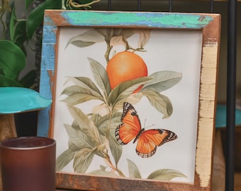 May include: A framed botanical print featuring an orange and a butterfly. The artwork is set in a rustic wooden frame with blue and green accents. A brown candle holder sits in the foreground.