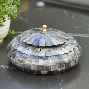 May include: A round decorative box with a lid, constructed from small, rectangular grey and blue tiles in a mosaic pattern. The lid features a scalloped edge and a small, gold-coloured knob. The box is likely for storage.