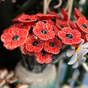 May include: A collection of ceramic poppy flowers with vibrant red petals and textured black centres, each mounted on a thin metal stem. The flowers are arranged in a cluster, showcasing a glossy finish and detailed petal ridges on each bloom.