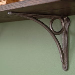 May include: A dark metallic shelf bracket with a curved support arm and a circular decorative element. The bracket is mounted to a wall and supports a dark wooden shelf. It features a textured, cast-metal finish with visible screw heads.