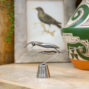 May include: A silver metal bird sculpture, crafted from spoons and other metal components. The bird has a detailed design and stands on a small, silver base. A framed bird print and a green and orange vase are visible in the background.