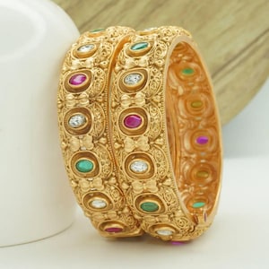 Puede incluir: Dos pulseras doradas con intrincados diseños en relieve. Cada pulsera presenta una serie de piedras preciosas de colores, incluyendo piedras rojas, verdes y transparentes. Las pulseras tienen una estética vintage, adecuadas para ocasiones especiales.