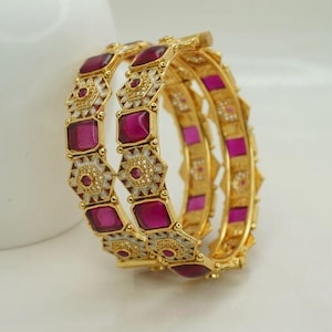Op de afbeelding: Gouden armbanden met een geometrisch ontwerp. Elke armband heeft afwisselende secties van magenta gekleurde stenen en wit en goud filigraan. De armbanden zijn gestapeld en tonen hun ingewikkelde details en levendige kleuren.