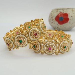 Puede incluir: Dos brazaletes dorados con detalles intrincados. Cada brazalete presenta diseños circulares con acentos blancos, rojos, verdes y dorados. Los brazaletes están adornados con pequeñas cuentas blancas redondas. Las joyas se exhiben sobre una superficie blanca.