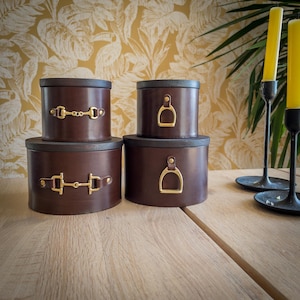 May include: Four brown leather storage boxes with dark wooden lids are stacked on a wooden surface. The boxes have gold-colored hardware, including stirrup and bit designs. Yellow candles in black candle holders are in the background.