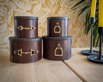 Puede incluir: Cuatro cajas de almacenamiento de cuero marrón con tapas de madera oscura apiladas sobre una superficie de madera. Las cajas tienen herrajes dorados, incluyendo diseños de estribos y bocados. Velas amarillas en candelabros negros en el fondo.