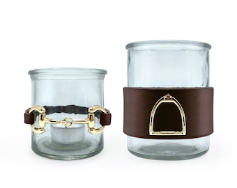 Puede incluir: Dos candelabros de cristal transparente. Uno tiene una correa de cuero marrón con un bocado de caballo dorado. El otro tiene una envoltura de cuero marrón con un diseño de estribo dorado. Sobre un fondo blanco.