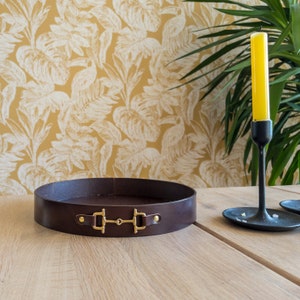 May include: A circular, dark brown leather tray with gold-coloured hardware. The tray features a horse bit design and sits on a light-coloured wooden table. A yellow candle in a black candlestick holder is to the right.