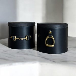 Könnte beinhalten: Zwei schwarze zylindrische Behälter mit Holzdeckeln. Einer hat ein goldfarbenes Gebiss-Design, der andere ein goldfarbenes Steigbügel-Design. Die Behälter stehen auf einer weißen Marmoroberfläche.