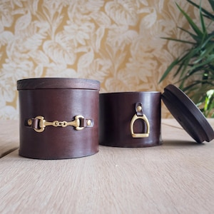 May include: Two brown leather containers with wooden lids. One container has a gold-colored horse bit design, and the other has a gold-colored stirrup design. The containers are cylindrical and sit on a light-colored wooden surface.