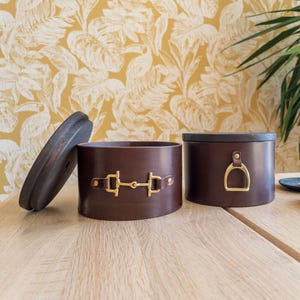 Op de afbeelding: Twee bruine leren opbergdozen met goudkleurige hardware. Eén doos heeft een deksel en de andere heeft een decoratief gespontwerp. De dozen zijn cilindrisch en staan op een houten oppervlak.