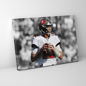 May include: A black and white photo of a football player wearing a white jersey with red accents and a black helmet with a skull and crossbones logo. The player is holding a football and looking to the right.