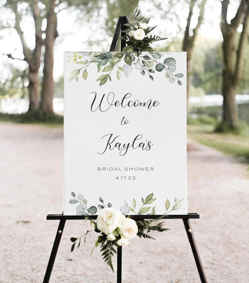 May include: White sign with green watercolor leaves and the text "Welcome to Kayla's Bridal Shower 4.17.23" on an easel.