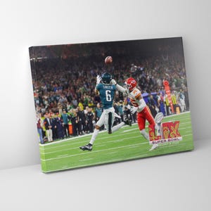 May include: A football player in a green and white uniform jumps to catch a football thrown by a player in a red uniform. The game is taking place in a stadium with a large crowd of spectators. The text "SUPER BOWL" is visible in the lower right corner of the image.