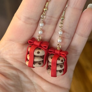 Puede incluir: Pendientes colgantes con pilas de mini galletas con chispas de chocolate, envueltas en cinta roja como regalos. Los pendientes tienen ganchos dorados y están adornados con una cadena de pequeñas perlas blancas.