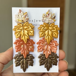 May include: A pair of dangle earrings featuring a layered leaf design in autumnal colors. The earrings showcase a gradient of colors, from light beige to gold, orange, and dark brown. The earrings are held against a white background.