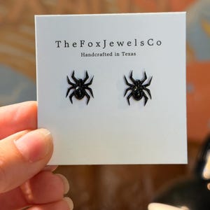 Op de afbeelding: Een paar zwarte spinvormige oorknopjes bevestigd op een witte displaykaart. De kaart bevat de tekst TheFoxJewelsCo en Handcrafted in Texas. De spinnen hebben een glanzende afwerking en acht afzonderlijke poten op de witte achtergrond.