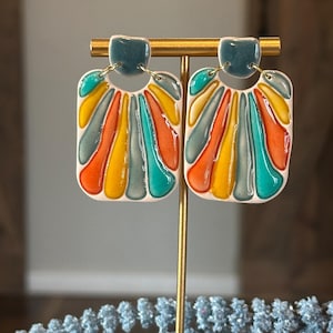 Puede incluir: Un par de pendientes de cerámica coloridos con un diseño geométrico. Los pendientes son una mezcla de turquesa, naranja, amarillo y blanco.