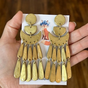 May include: A pair of gold-toned dangle earrings with a crescent moon shape and multiple teardrop-shaped charms. The earrings are displayed on a white card with a fox illustration.