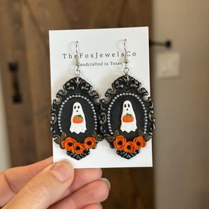 Op de afbeelding: Zwart omrande Halloween-oorbellen met een witte geest die een oranje pompoen vasthoudt. De oorbellen hebben oranje bloemen en zijn handgemaakt in Texas. The Fox Jewels Co.