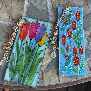 Peut inclure: Deux marque-pages rectangulaires au style vitrail. L'un présente des tulipes rouges, roses, violettes et jaunes avec des tiges vertes. L'autre montre des tulipes rouges et des colibris sur fond bleu. Les deux ont des chaînes dorées avec breloques.
