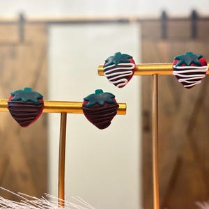 May include: A pair of earrings shaped like strawberries dipped in chocolate and white frosting. The earrings are displayed on a gold stand.