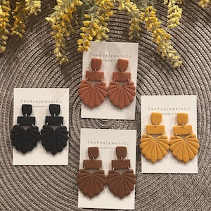 Pode incluir: Quatro pares de brincos de argila polimérica feitos à mão nas cores preto, ferrugem, castanho e amarelo mostarda. Cada par apresenta uma parte superior retangular, uma seção do meio e uma parte inferior em forma de leque. Os brincos são exibidos em cartões brancos com "The Fox Jewels Co."